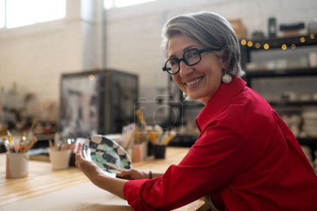 Téléchargez les photos : Potier femme âgée positive dans un tablier posant dans un atelier de céramique, tenant une plaque artisanale, regardant la caméra et souriant. - en image libre de droit