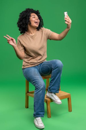 Foto de Conectado. Mujer joven emocionada sentada en la silla con un teléfono en las manos - Imagen libre de derechos