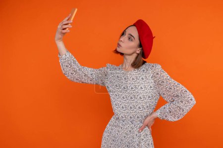 Téléchargez les photos : Femme adulte romantique portant une robe élégante et un béret rouge debout faisant selfie envoyant de l'air embrasser isolé sur fond orange. - en image libre de droit