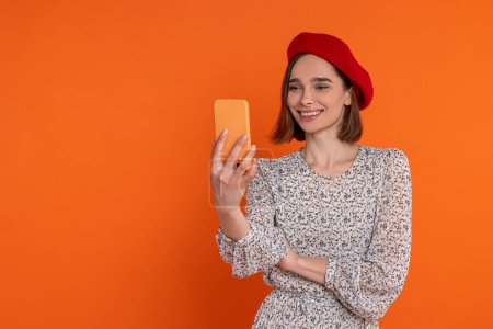 Téléchargez les photos : Belle femme adulte portant une robe élégante et béret rouge faisant selfie sur téléphone portable debout isolé sur fond orange. - en image libre de droit