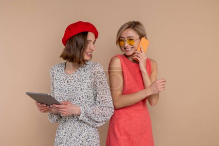 Téléchargez les photos : Deux amies portant des robes élégantes se tenant ensemble isolées sur fond beige en utilisant une tablette numérique et un téléphone portable. - en image libre de droit
