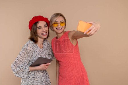 Foto de Dos amigas con vestidos elegantes de pie juntas aisladas sobre fondo beige usando tableta digital y teléfono móvil. - Imagen libre de derechos