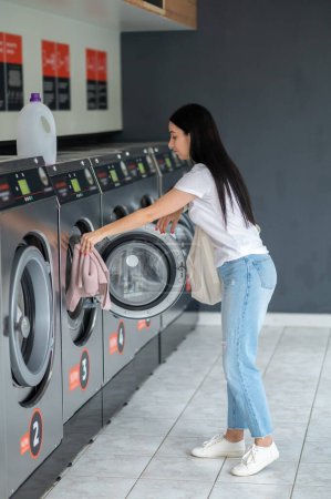 Téléchargez les photos : Brunette femme insérant des vêtements sales dans la machine à laver dans la buanderie. - en image libre de droit