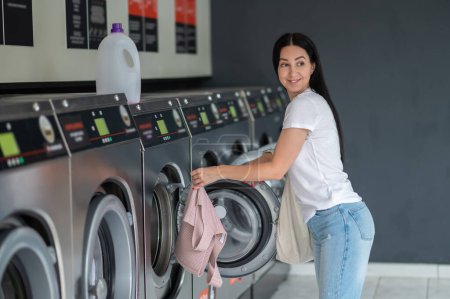 Téléchargez les photos : Brunette femme insérant des vêtements sales dans la machine à laver dans la buanderie. - en image libre de droit