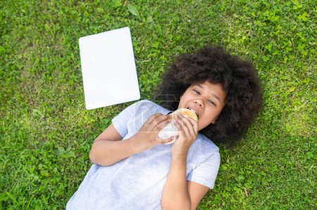 Photo for African dark skinned girl lying on grass and eating fast food in park, happy kid having snack after using laptop outdoor. - Royalty Free Image
