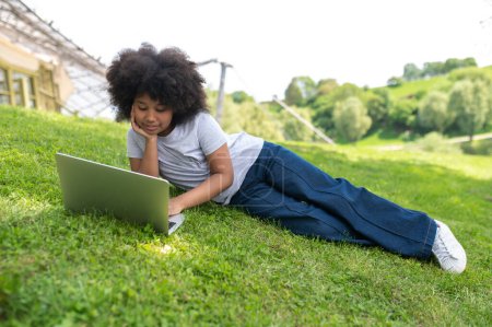 Photo for Charming little girl with curly hair using laptop in summer garden having video call watching movie online lesson distance education. - Royalty Free Image