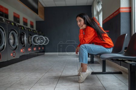Foto de Mujer aburrida y cansada con capucha naranja sentada en la lavandería automática mientras espera a que se haga la colada. - Imagen libre de derechos