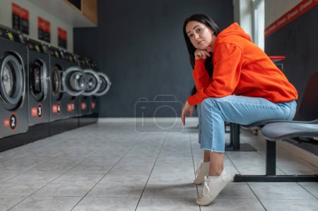 Photo for Tired bored woman wearing orange hoodie sitting in automatic laundry room while waiting for the laundry to be done. - Royalty Free Image
