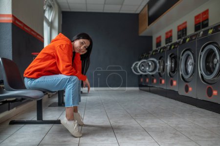 Müde gelangweilte Frau mit orangefarbenem Kapuzenpullover sitzt in der automatischen Waschküche und wartet auf die Wäsche.