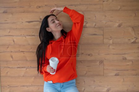 Téléchargez les photos : Femme optimiste en sweat à capuche orange élégant avec boisson chaude à emporter debout regardant loin près du mur en bois. - en image libre de droit