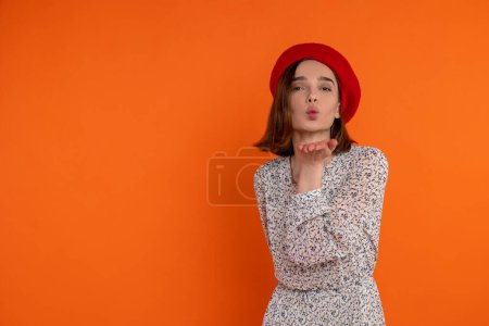Téléchargez les photos : Femme adulte romantique portant une robe élégante et un béret rouge envoyant de l'air s'embrasser debout isolé sur fond orange. - en image libre de droit