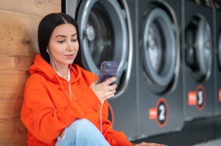 Foto de Young woman wearing orange hoodie listening music with smartphone and earphones waiting in laundry room. - Imagen libre de derechos