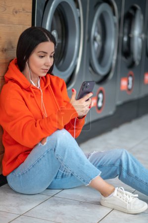 Téléchargez les photos : Young woman wearing orange hoodie listening music with smartphone and earphones waiting in laundry room. - en image libre de droit
