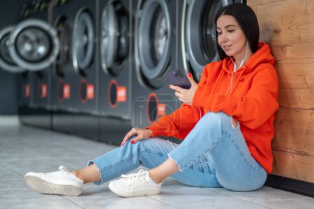 Photo for Young woman wearing orange hoodie listening music with smartphone and earphones waiting in laundry room. - Royalty Free Image