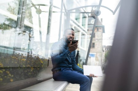 Foto de Hombre afroamericano. Barbudo joven afroamericano hombre sentado en el banco con un teléfono en las manos - Imagen libre de derechos