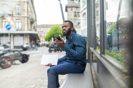 Foto de Hombre afroamericano. Barbudo joven afroamericano hombre sentado en el banco con un teléfono en las manos - Imagen libre de derechos