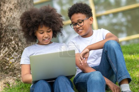 Téléchargez les photos : Petits enfants amis fille et garçon avec ordinateur portable assis sur l'herbe près de l'arbre regarder film navigation sur Internet. - en image libre de droit