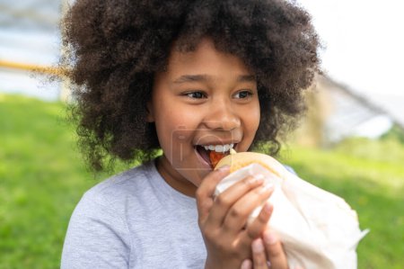 Foto de Niña de piel oscura comiendo comida chatarra con placer en el parque mientras está sentada sobre hierba verde, niña con peinado afro usando ropa casual. - Imagen libre de derechos