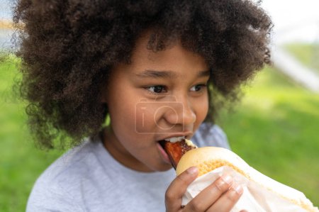 Photo for Dark skinned female child eating junk food with pleasure in park while sitting on green grass, girl with afro hairstyle wearing casual clothing. - Royalty Free Image