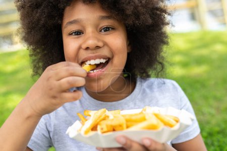 Photo for Dark skinned female child eating junk food with pleasure in park while sitting on green grass, girl with afro hairstyle wearing casual clothing. - Royalty Free Image