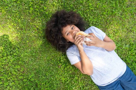 Photo for African dark skinned girl lying on grass and eating fast food in park, happy kid having snack after using laptop outdoor. - Royalty Free Image