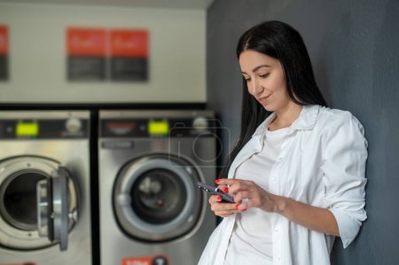 Photo for Young woman waiting in a laundry room using mobile phone, browsing internet. - Royalty Free Image