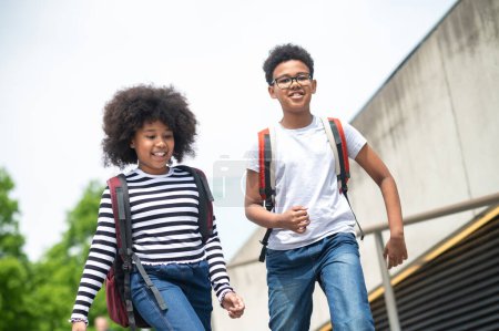 Téléchargez les photos : Écoliers actifs courant dans les escaliers avec des sacs à dos portant des chemises décontractées et des jeans marchant en s'amusant après l'école. - en image libre de droit