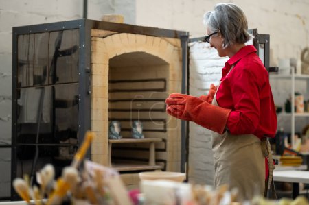 Foto de Madura artesana tomando productos de arcilla del horno de cerámica, trabajo a mano en taller de cerámica. - Imagen libre de derechos