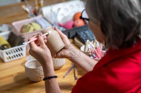Téléchargez les photos : Femme mature méconnaissable peignant sur terre cuite dans un atelier de poterie, décorant des produits en argile. - en image libre de droit