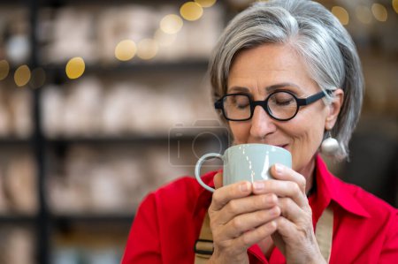 Photo for Satisfied senior craftswoman resting at pottery workshop drinking coffee smelling aromat. - Royalty Free Image