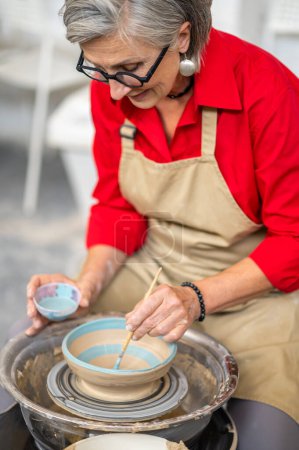 Photo for Mature craftswoman painting clay mug blue paint, using pottery wheel and paintbrush, creating earthenware in workshop. - Royalty Free Image
