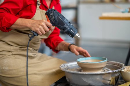 Téléchargez les photos : Femme méconnaissable maître en tablier séchage sculpture en argile avec pistolet à chaleur dans l'atelier de poterie. - en image libre de droit