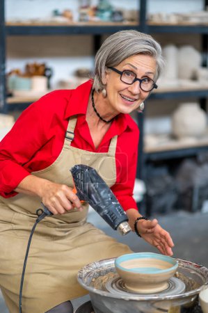 Foto de Senior master mujer en delantal secado escultura de arcilla con pistola de calor en taller de cerámica. - Imagen libre de derechos