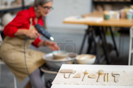 Téléchargez les photos : Equipements de poterie avec maître femme senior en tablier séchage sculpture en argile avec pistolet à chaleur dans l'atelier de poterie. - en image libre de droit