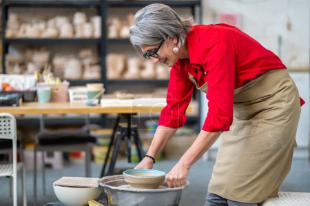 Photo for Talented senior woman potter taking finished plate from pottery wheel, making earthenware. - Royalty Free Image