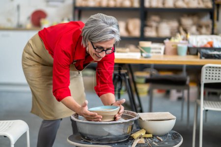 Talented senior woman potter taking finished plate from pottery wheel, making earthenware.