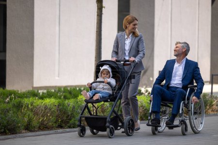 Téléchargez les photos : Parents marchant dans le parc, homme en fauteuil roulant, femme avec poussette bébé, loisirs en famille. - en image libre de droit