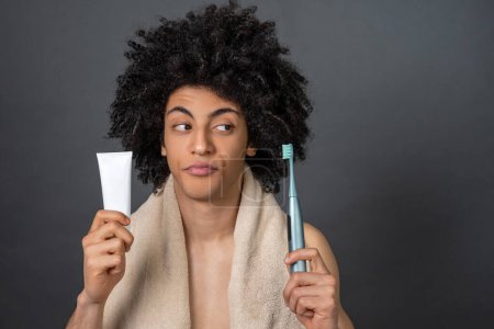 Téléchargez les photos : Le choix. Jeune homme tenant brosse à dents et dentifrice et l'air incertain - en image libre de droit