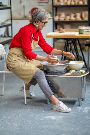 Téléchargez les photos : Femme mûre travaillant sur la roue des potiers créant des produits en argile à son atelier. - en image libre de droit