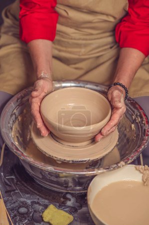 Photo for Hands of unrecognizable mature woman master of ceramics working on a potters wheel, making plate of clay in art studio. - Royalty Free Image