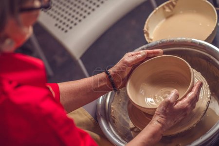 Foto de Manos de mujer madura irreconocible maestra de la cerámica trabajando en una rueda de alfarero, haciendo plato de arcilla en el estudio de arte. - Imagen libre de derechos