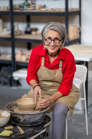 Téléchargez les photos : Femme mûre travaillant sur la roue des potiers créant des produits en argile à son atelier. - en image libre de droit