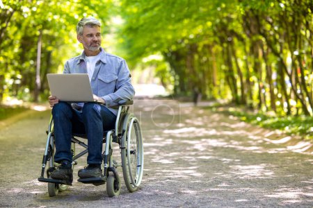 Téléchargez les photos : Homme aux cheveux gris freelance avec handicap en fauteuil roulant portant des vêtements décontractés travaillant en ligne. - en image libre de droit
