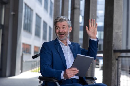 Téléchargez les photos : Heureux homme d'affaires amical en fauteuil roulant avec tablette PC dans les mains, navigation sur le web, en utilisant un appareil mobile pour travailler à l'extérieur, saluant quelqu'un avec la main agitant. - en image libre de droit
