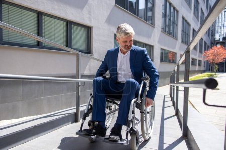 Photo for Adult Caucasian gray haired businessman in formal wear gets to work in wheelchair rides ramp, disabled person. - Royalty Free Image