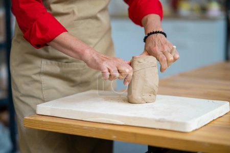 Téléchargez les photos : Technique de pétrissage manuel en poterie. Femme méconnaissable argile de coupe avec fil debout derrière la table en studio. - en image libre de droit