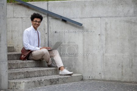 Photo for Working remotely. Curly-haired young man in white shirt sitting on the steps with laptop - Royalty Free Image