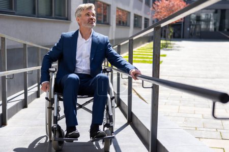 Adult Caucasian gray haired businessman in formal wear gets to work in wheelchair rides ramp, disabled person.