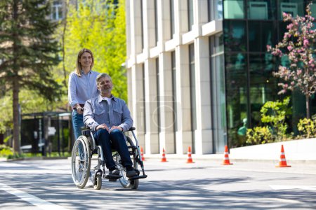 Téléchargez les photos : Mari heureux en fauteuil roulant et femme aimante marchant dans la rue de la ville. - en image libre de droit