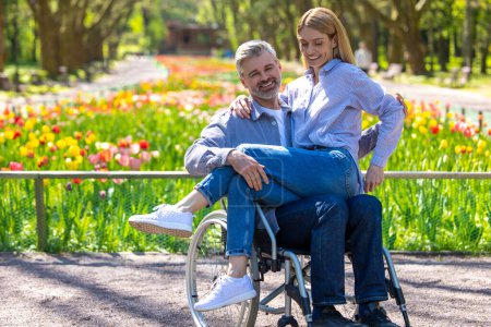 Foto de Familia feliz descansando en el parque, mujer y hombre discapacitado en silla de ruedas,, pareja abrazándose. - Imagen libre de derechos